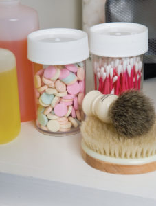 An 8oz Storage Jar sits among cotton swab containers, colorful tablets, bottles of liquid, and nearby shaving and body brushes.