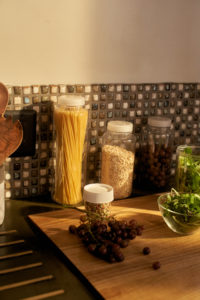 Jars of pasta, oats, and beans with a 16oz storage bottle on the counter next to grapes and greens on a wooden cutting board.