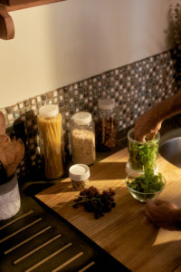 A person prepares fresh greens on a wooden kitchen counter near a 4oz Storage Jar filled with pasta, along with oats, hazelnuts, and grapes.