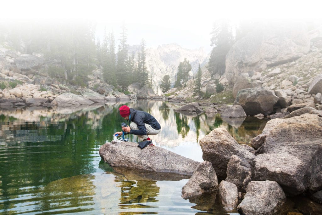 Person in a red beanie crouches on a rock by a calm mountain lake surrounded by trees and rocky cliffs.