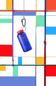 A 32oz Wide Mouth Sustain Water Bottle in blue with an orange cap hangs from a carabiner among colorful ropes and geometric shapes.
