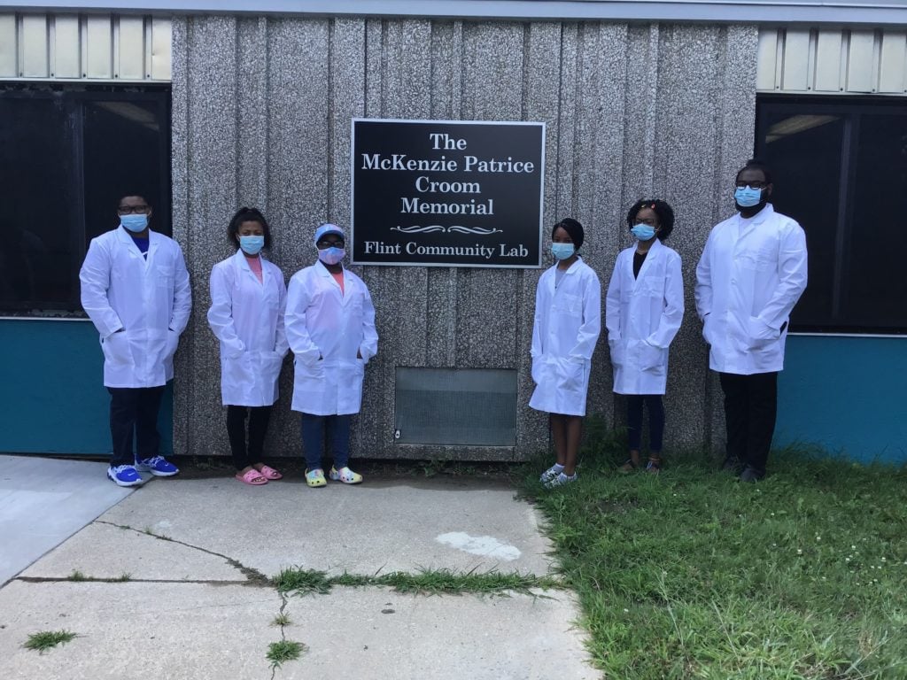 Six people in lab coats and masks stand outside the Flint Community Lab under a memorial sign.