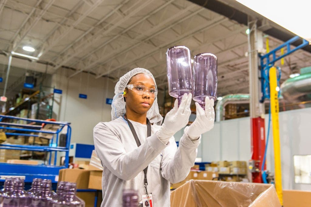 A worker inspects two purple plastic bottles in a factory wearing gloves, a hairnet, and safety glasses.