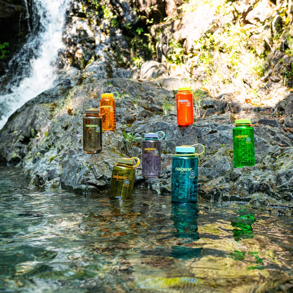 Six colorful water bottles sit on rocks by a waterfall, with clear water in the foreground and greenery around.