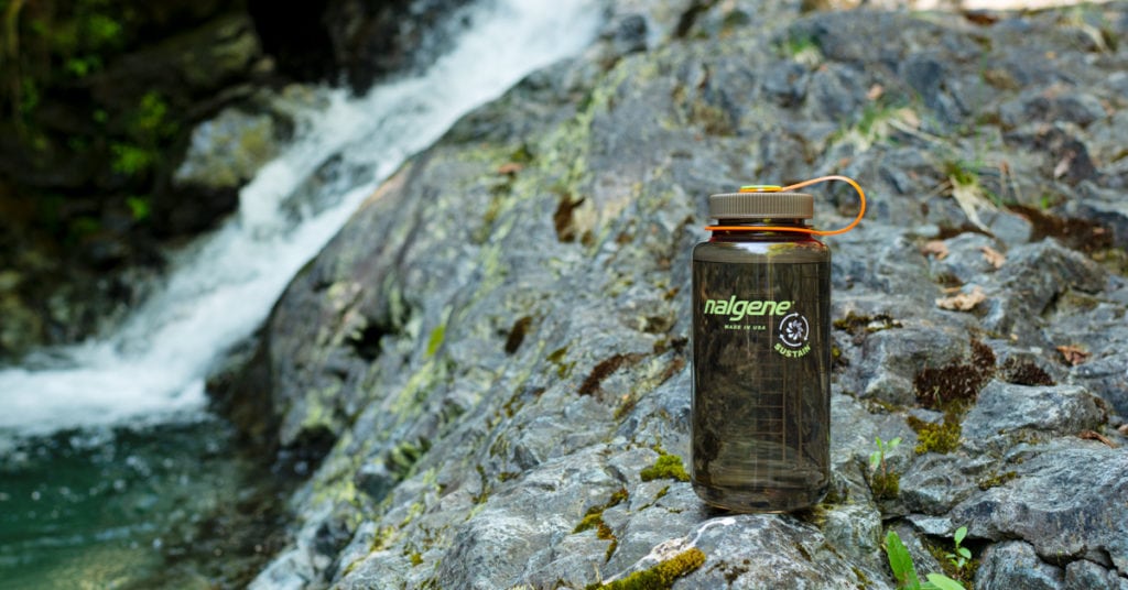 A Nalgene water bottle rests on a rocky surface beside a flowing waterfall and pool.