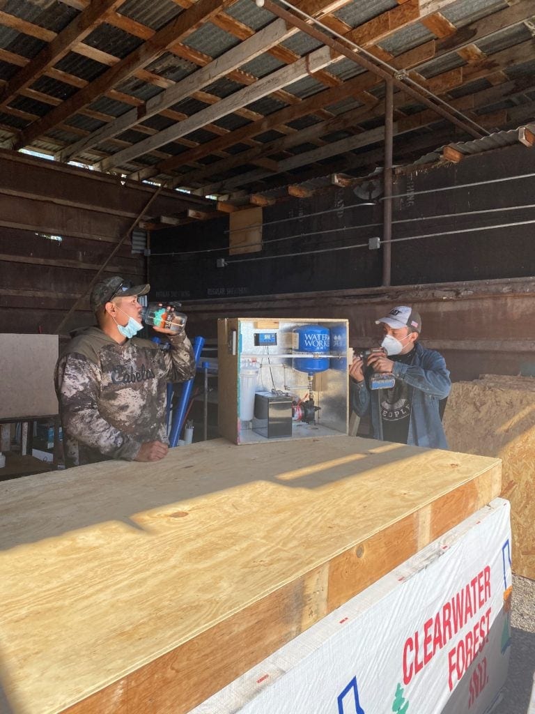 Two people wearing masks drink from cups inside a rustic building near a water station and a Clearwater Forest sign.