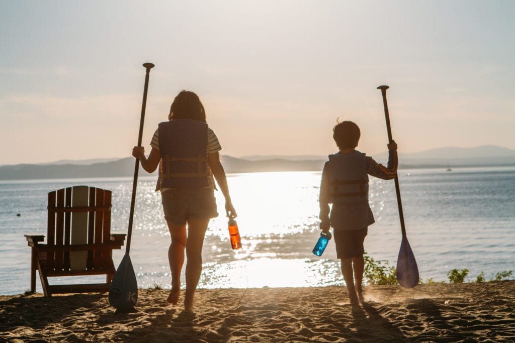 Two kids with life jackets and paddles walk toward a lake at sunset, holding bottles, seen from behind.