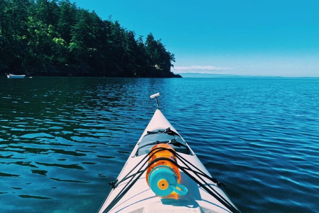 View from a kayak on calm blue water near a forested shoreline under a clear sky.