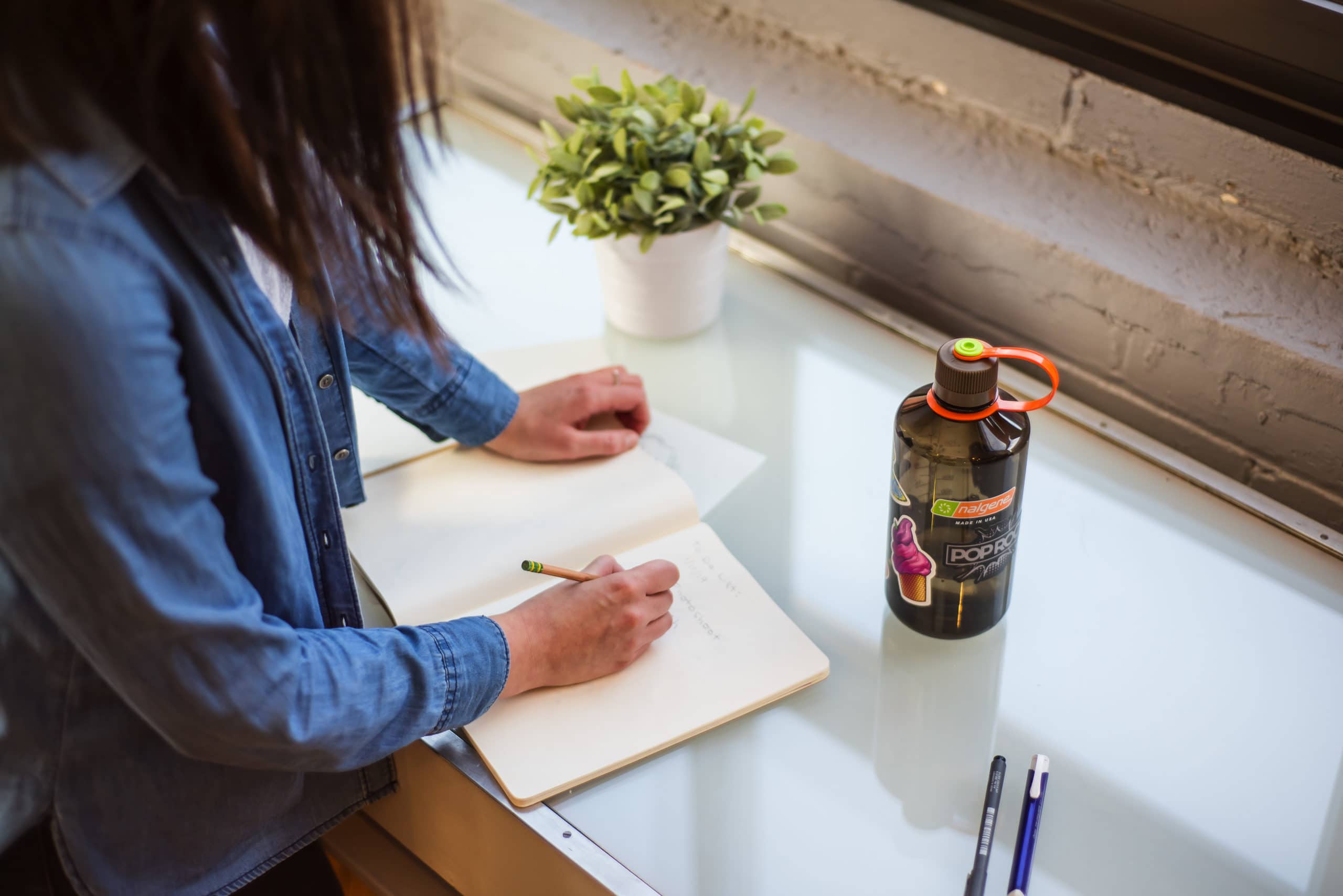 Person writing in a notebook at a desk with a plant, water bottle, and pens nearby.