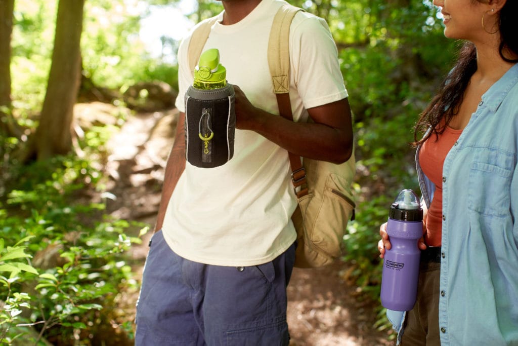 Two people holding water bottles while hiking on a forest trail, surrounded by greenery.