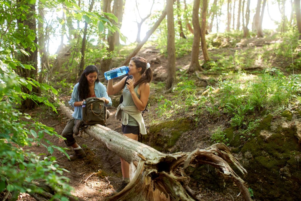 Two women hiking in a forest; one sits on a log, the other drinks water and smiles in the sunlight.