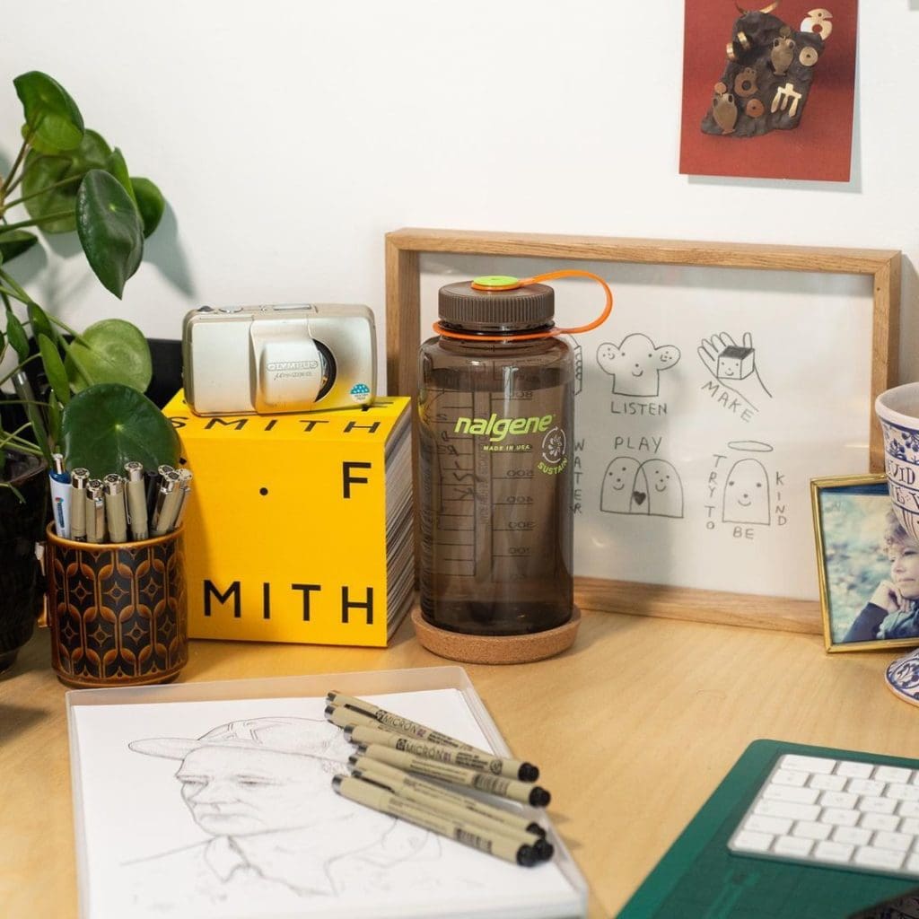 A desk with pens, a sketch, a Nalgene bottle, plants, art, a mug, a keyboard, and a framed drawing.