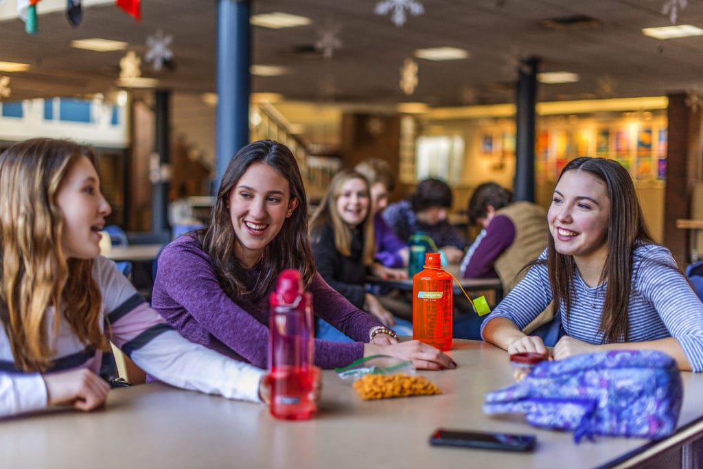 Three teenage girls sit at a table in a cafeteria, talking and smiling with snacks and drinks in front of them.
