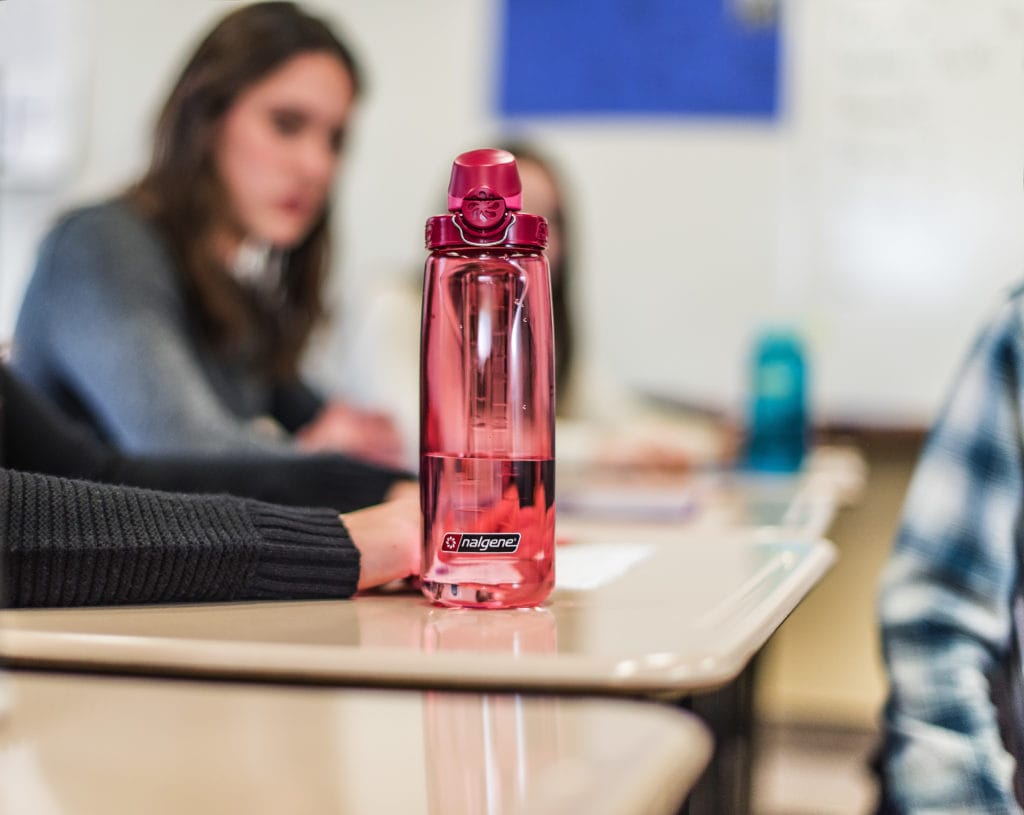 A pink Nalgene water bottle on a classroom desk with blurred students in the background.