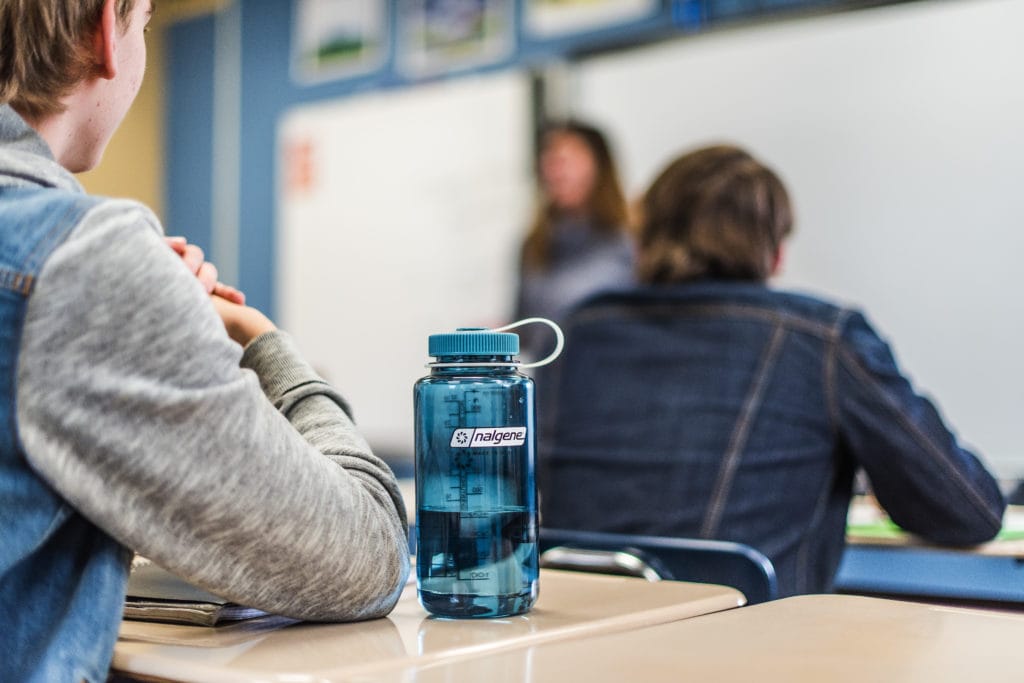 A blue water bottle on a student's desk in a classroom with students and a teacher in the background.