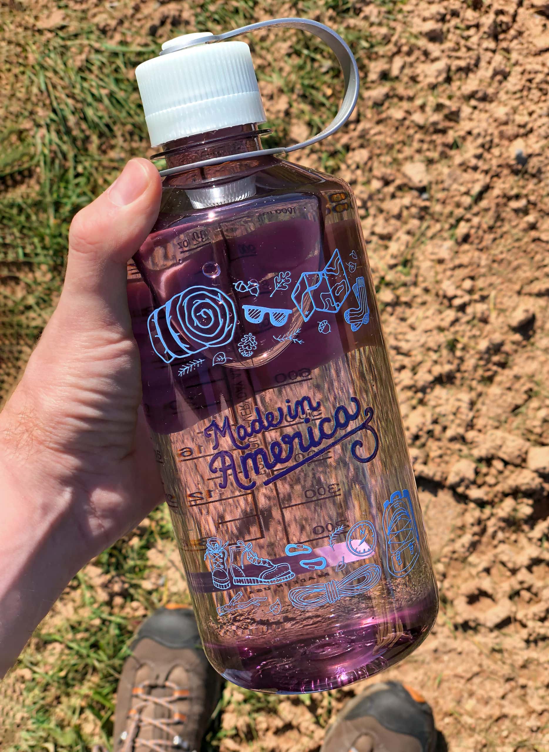 Hand holding a clear purple water bottle labeled "Made in America" over dirt and grass, with boots visible below.