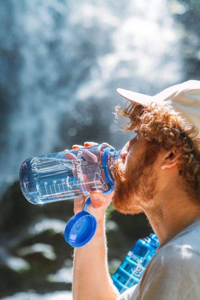 A man with curly hair drinks water from a large blue bottle outdoors near a waterfall.