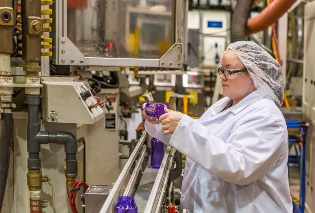 A worker in a lab coat and hairnet inspects purple plastic bottles on a factory production line.