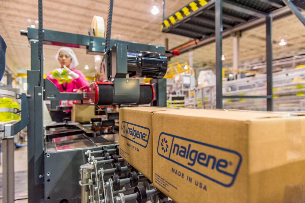 Cardboard boxes labeled "nalgene" move on a conveyor belt in a warehouse; worker in background.
