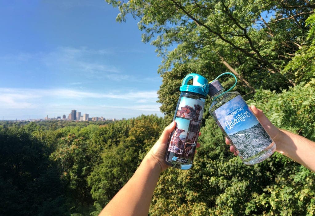 Two hands hold water bottles up in front of a city skyline and lush green trees under a clear blue sky.