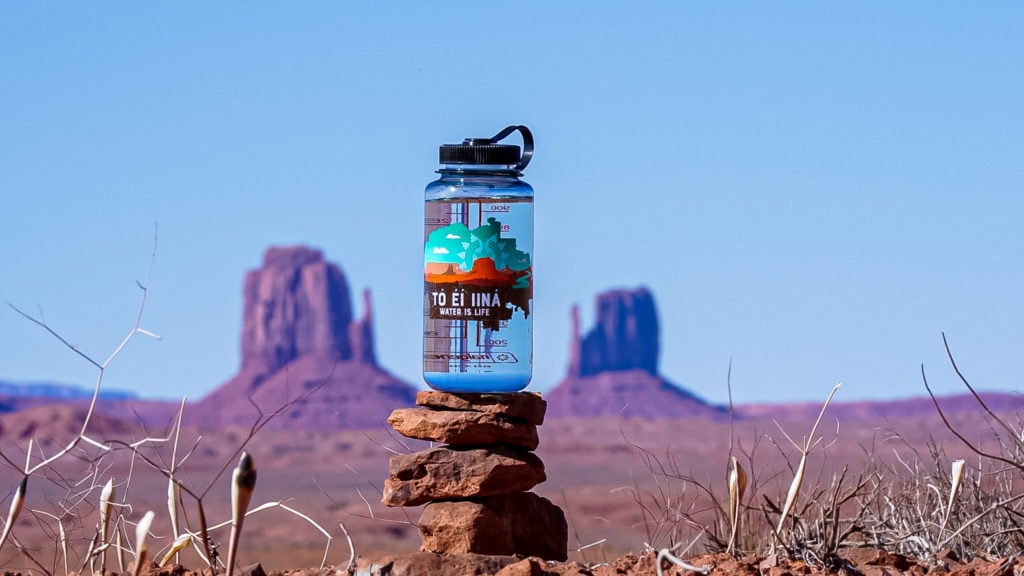 A water bottle on stacked rocks in the desert, with Monument Valley buttes in the background under a clear sky.