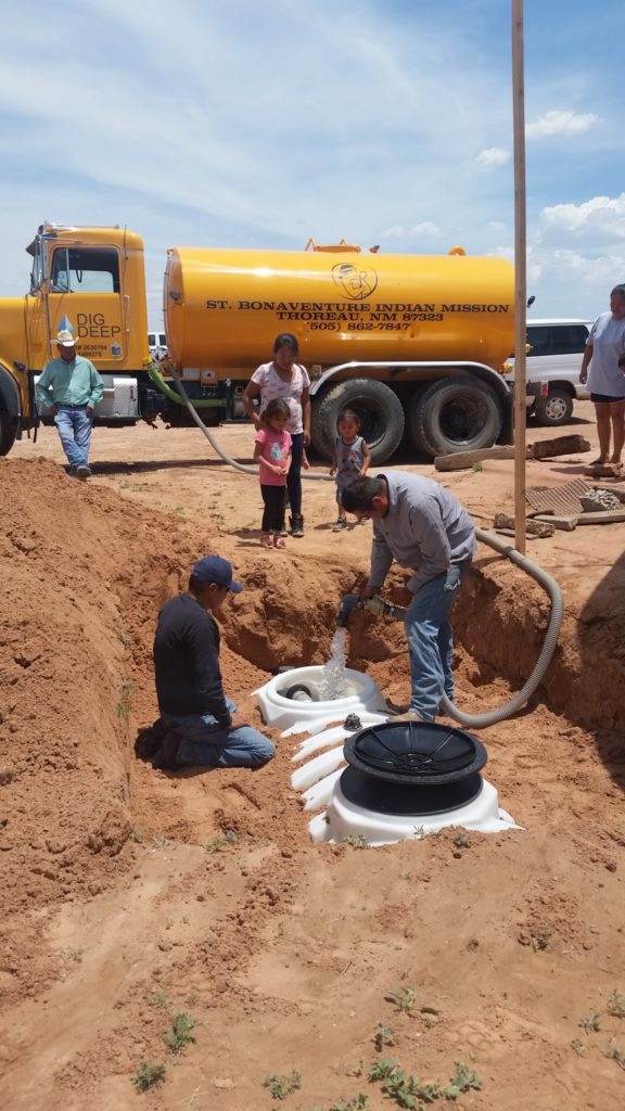Two men install a water system in a dug pit while others watch; a water truck is parked nearby.