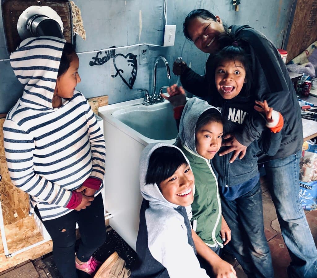 Four smiling children and an adult gather around a sink in a cozy, rustic indoor setting.