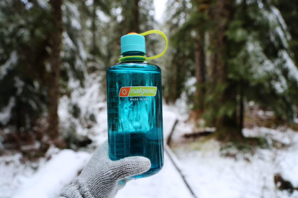A gloved hand holds a blue Nalgene water bottle in a snowy forest.