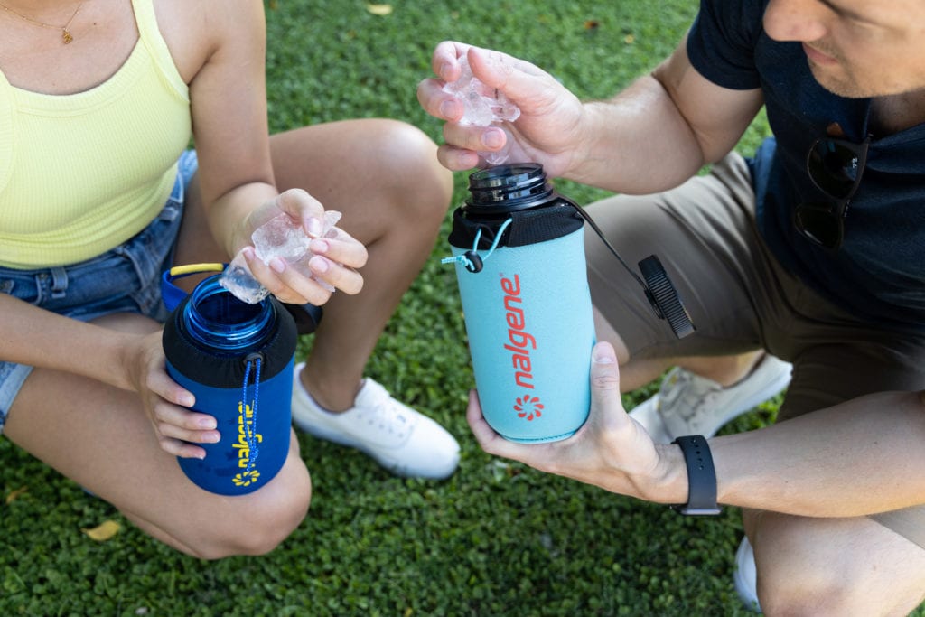 Two people holding Nalgene bottles and adding ice cubes, sitting on grass outdoors.