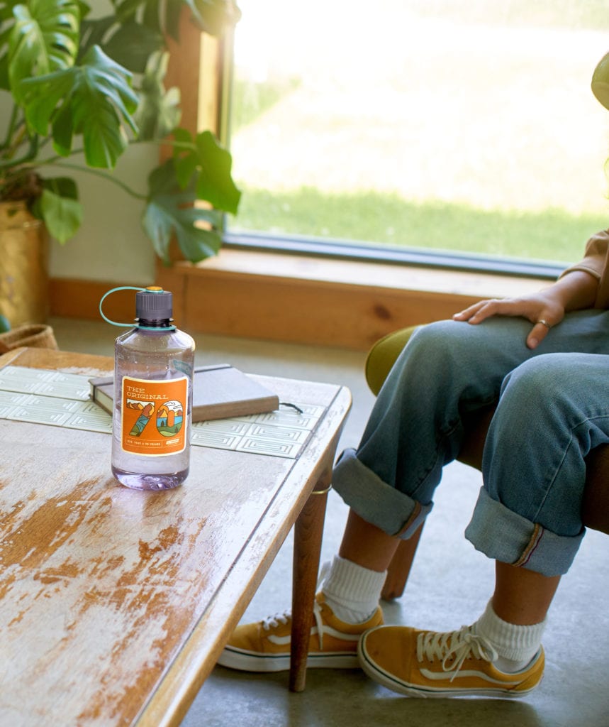 A water bottle on a table next to a seated person in rolled-up jeans and yellow sneakers.