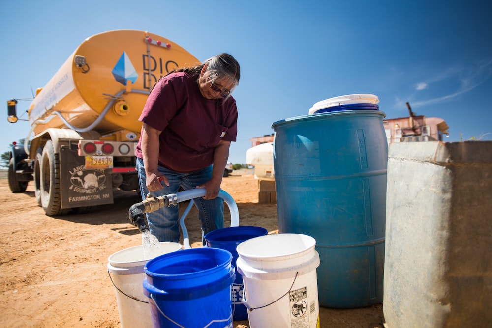 A person fills white and blue buckets with water from a hose, near a large water truck on a sunny day.
