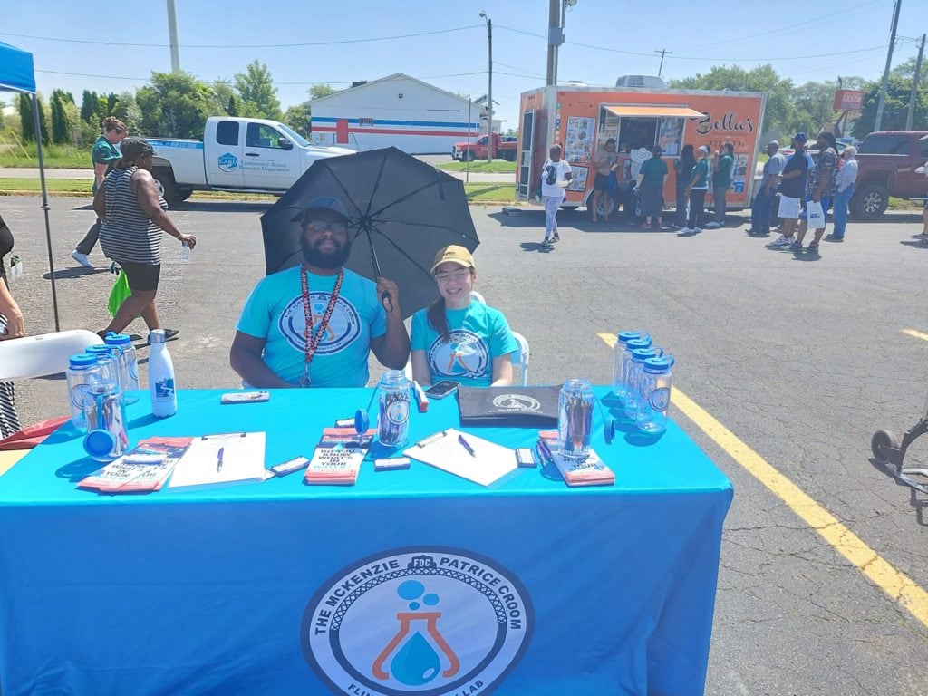 Two people sit at an outdoor info booth with blue tablecloth; crowd and food truck in background.