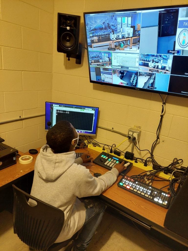 A person operates video and audio controls in a media production room with multiple screens and equipment.