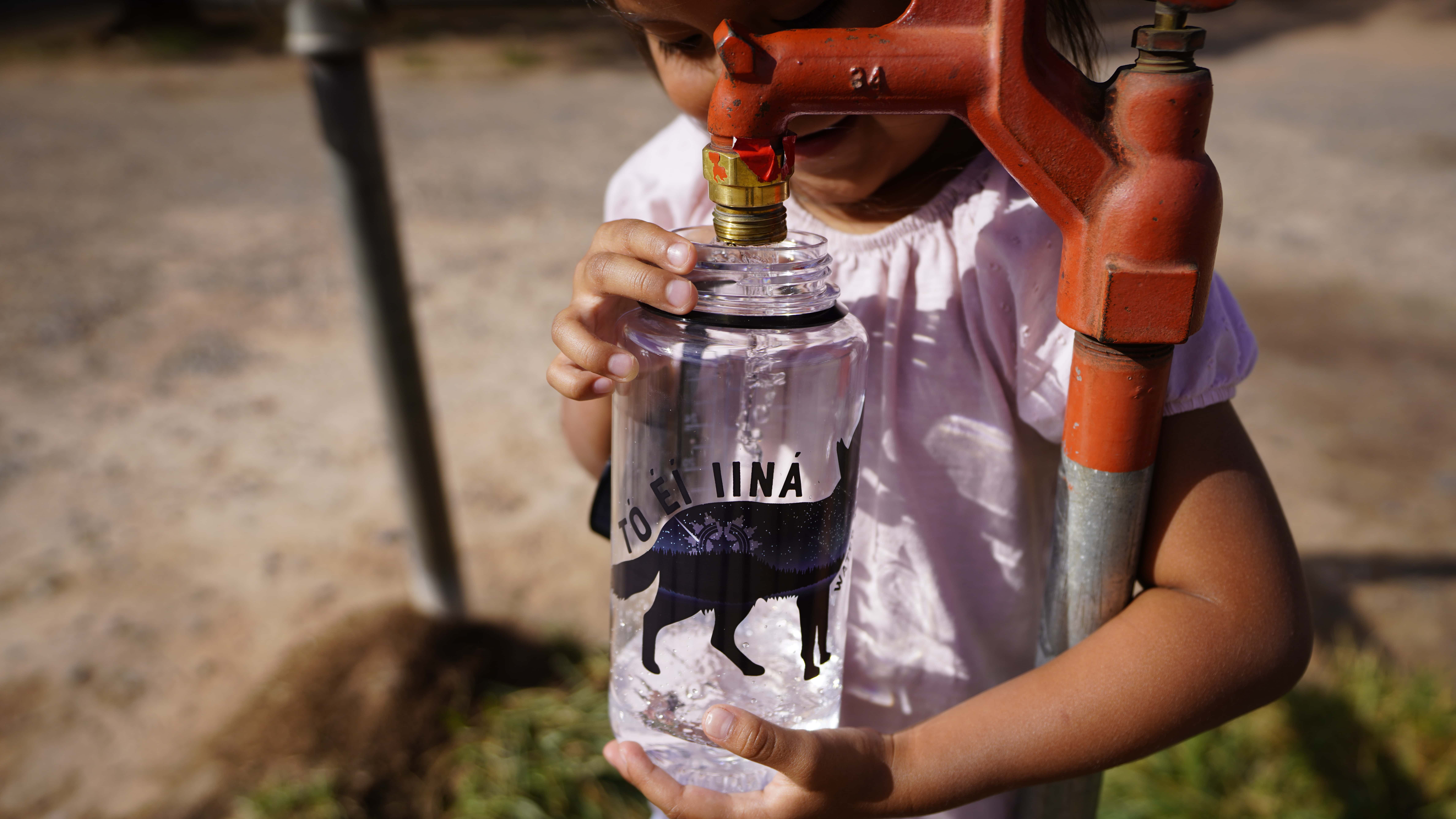 A child fills a water bottle from an outdoor red faucet on a sunny day.