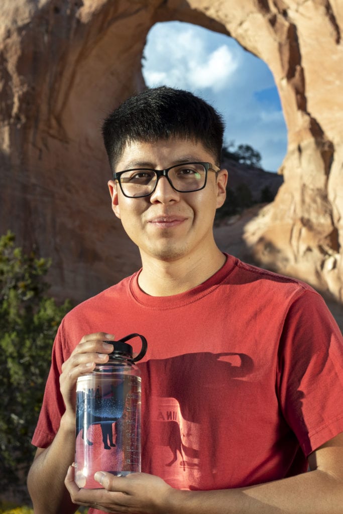 A man in glasses holds a water bottle, standing in front of a large rock arch under a blue sky.