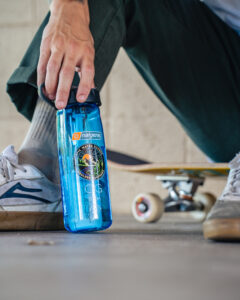 Hand holding a 32oz/24oz Epic Filter Cap + Kit next to a skateboard and white sneakers on a concrete floor.