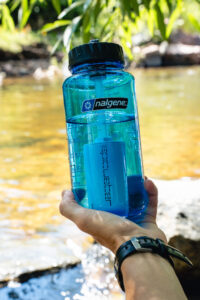 A hand holds a blue 32oz/24oz Epic Filter Cap + Kit water bottle beside a clear stream outdoors.