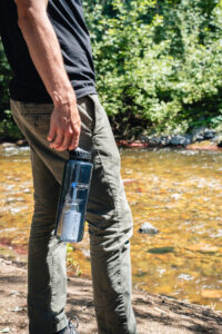 Person holding a 48oz Epic Filter Cap + Kit bottle by a river, outdoors in sunlight with greenery in the background.