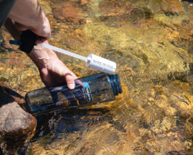 A person fills a 48oz Epic Filter Cap + Kit bottle from a clear, shallow stream.