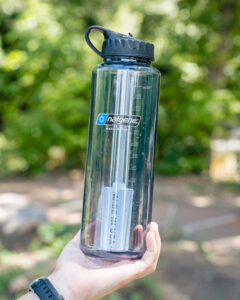 A hand holds a 48oz Epic Filter Cap + Kit water bottle outdoors with trees softly blurred in the background.