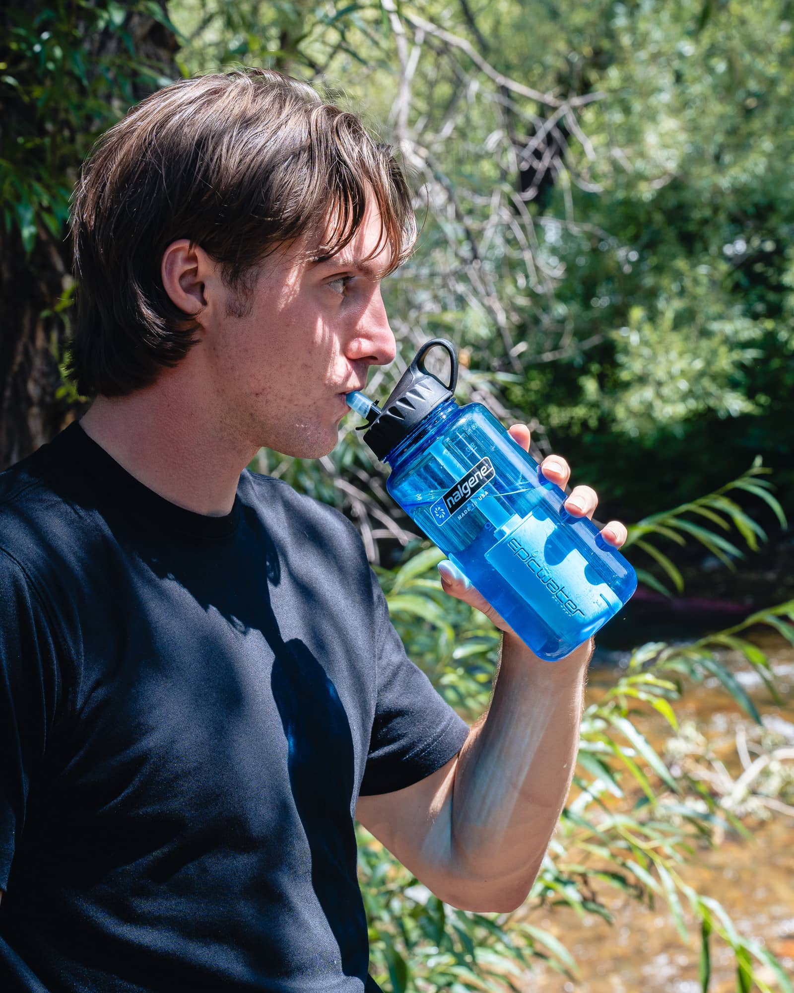 A man in a black shirt drinks water from a blue bottle outdoors near trees and a stream.