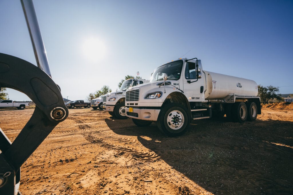 White tanker trucks parked on a sunny, dirt lot with tire tracks and equipment visible in the foreground.