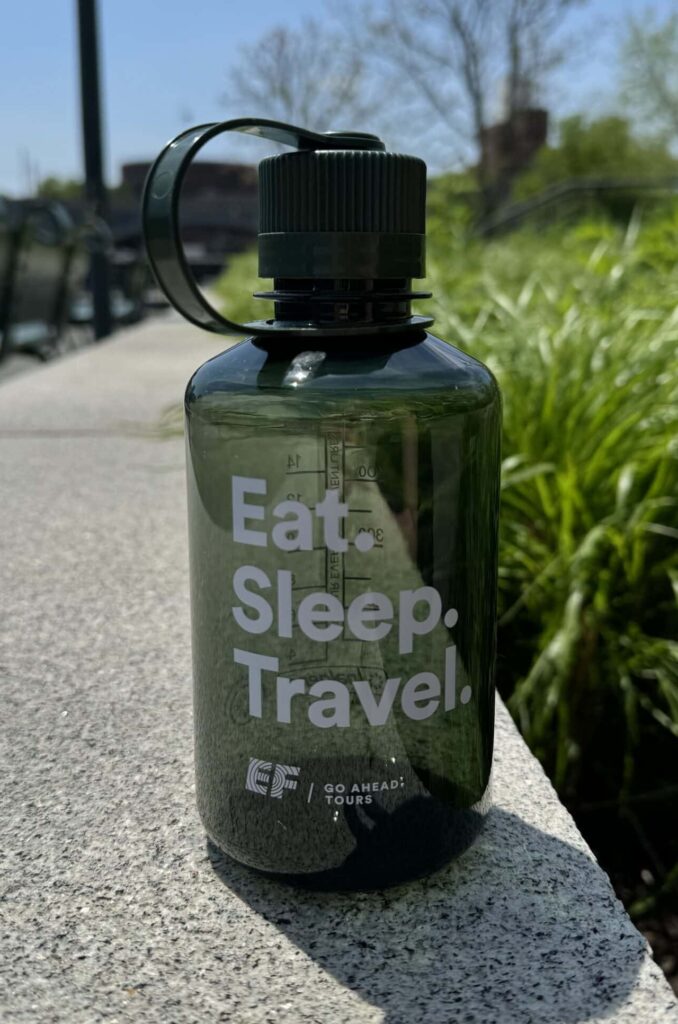 A dark green water bottle with "Eat. Sleep. Travel." written on it, sitting on a stone ledge outdoors.