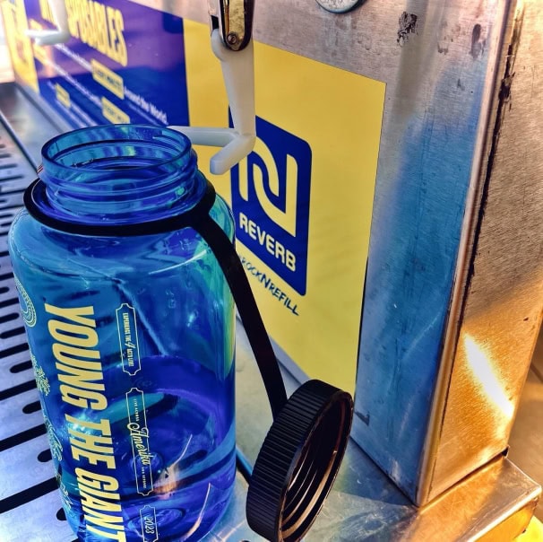 A blue water bottle being filled at a public refill station, with its lid hanging to the side.