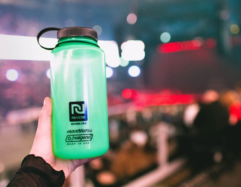 A hand holds a green reusable Nalgene water bottle at an indoor event with colorful lights.
