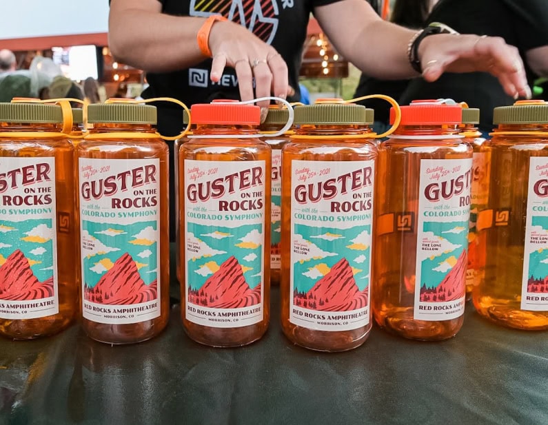 Six Nalgene bottles with "Guster on the Rocks" labels lined up on a table; hands reach in from above.
