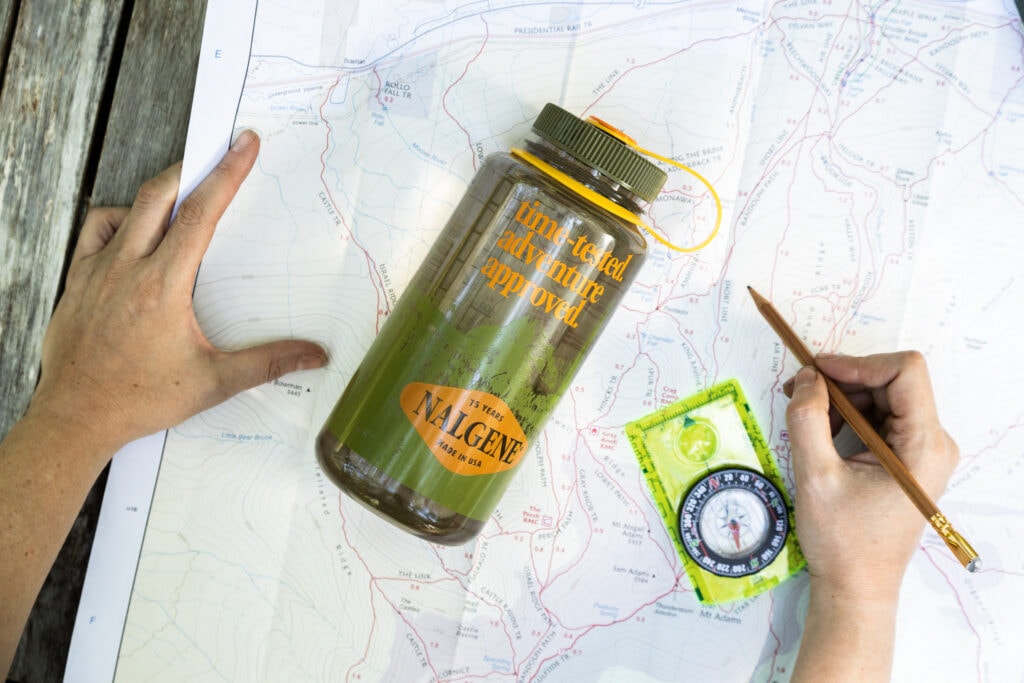 A hand draws on a map with a pencil beside a green compass and a Nalgene water bottle.