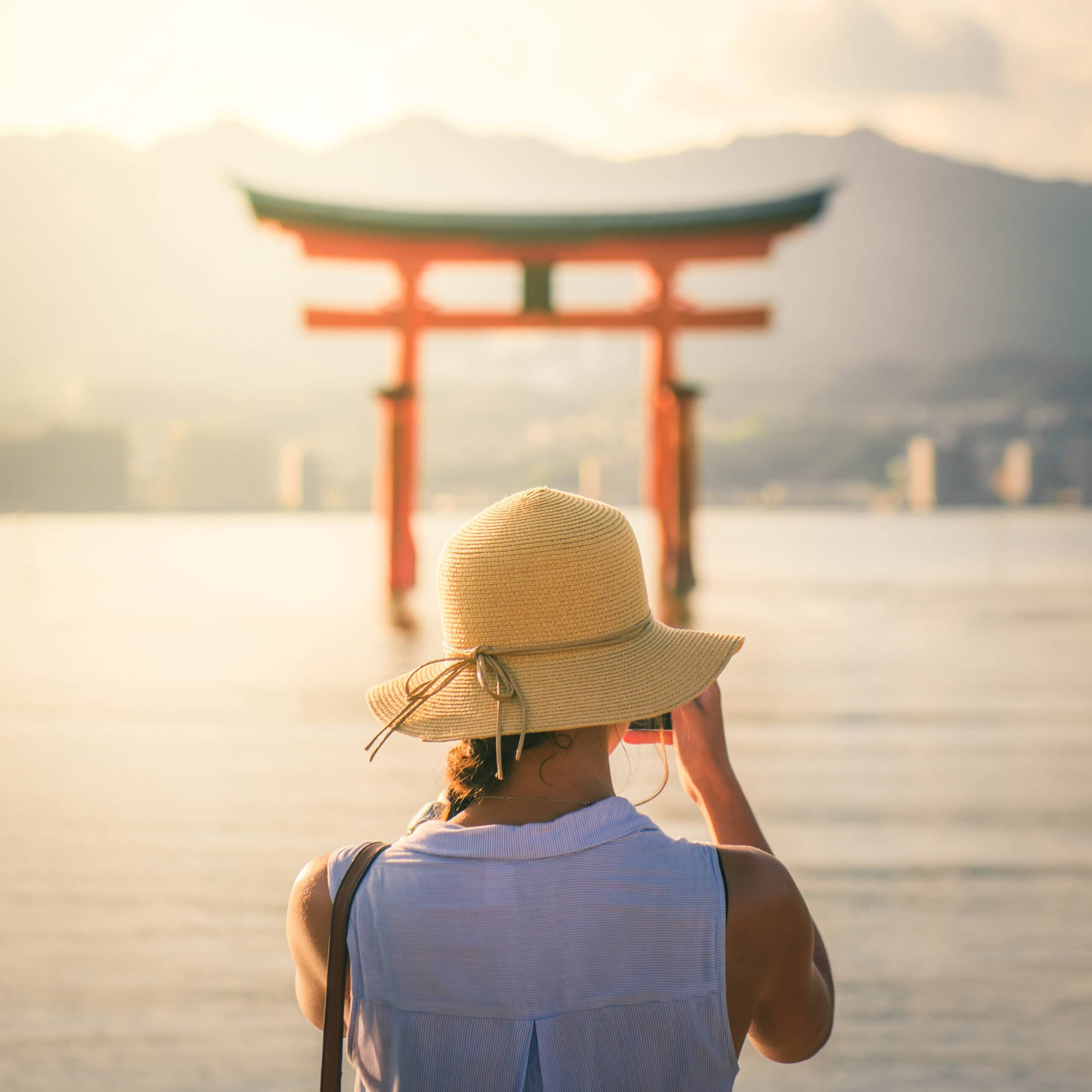 A person in a sunhat takes a photo of a red torii gate over water at sunset.