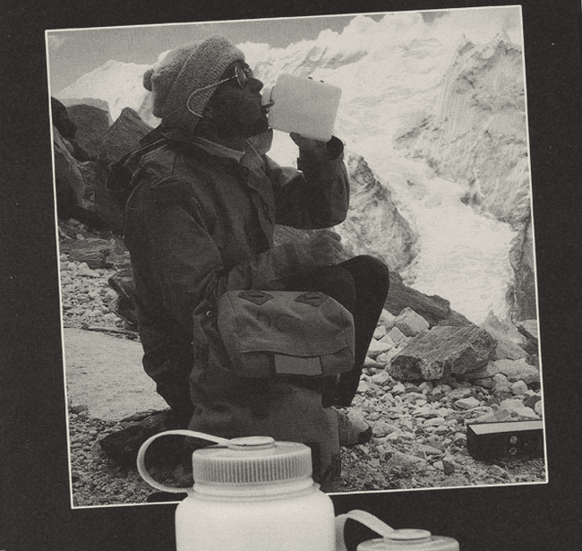A person in winter gear drinks from a bottle while sitting on rocky terrain near snowy mountains.