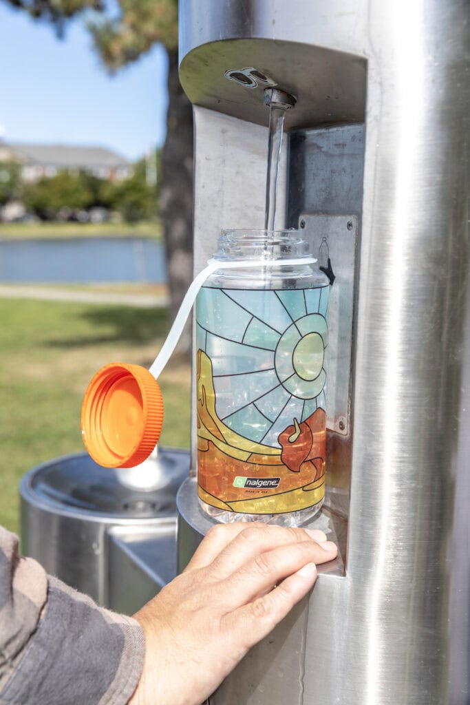 A water bottle being filled at an outdoor water fountain, with a hand holding the bottle in place.
